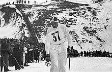 A man in white is pictured with a ski pole in his left hand. On his shirt is pinned a number tag with the number 37. Behind him to his right is a crowd of spectators at the side of a cross-country skiing course.