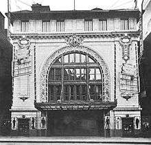 Black and white photo of a theater with a vaulted ceiling and an ornate curtain in front of the stage.