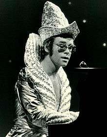 An image of Elton John playing the piano while wearing an elaborate outfit.