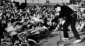 A dark-haired man wearing dark clothing, performing on stage to a crowd, many of whom are stretching out their arms towards him