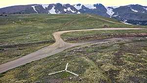 An aerial view of the emergency backcountry helicopter landing zone located at the summit of Rollins Pass; the white prong of the landing zone, furthest from the camera, points north. The comparatively low saddle of Rollins Pass is visible in this image as the summit itself (in the foreground and midground) is plainly lower in elevation than the surrounding mountains. The wood debris consists of both remnants from snowsheds that covered the tracks as well as discarded railroad ties that were removed from service in the summer of 1936.