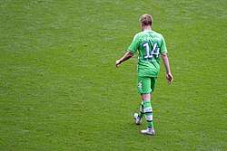 A colour photograph of Kevin de Bruyne, face back to the camera, wearing an all-green kit.