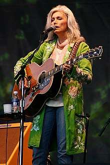 A woman with long grey hair wearing a long green jacket, playing a guitar and singing into a microphone