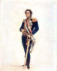 Colored full-length portrait showing a young man with curly hair and long sideburns who is wearing an elaborate gold-embroidered blue military tunic with gold epaulets and medals, blue trousers, black boots, a striped sash of office and gold belt, with his left hand resting on a sheathed sword