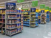 Aisles in a supermarket with small sets of shelves on the end, receding from left to right. Atop each set of shelves is a large white on black sign with the price of an item for sale on those shelves. White on green signs with the types of products in the aisles hang from the high ceiling.