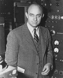 A man looking to the camera, wearing a suit. He is standing in front of a machine.