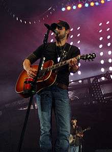 A man wearing a baseball cap, sunglasses and dark clothing, playing a guitar and singing into a microphone