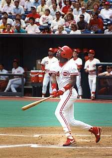 A dark-skinned young man wearing a white baseball uniform with red trim and a red batting helmet swinging a baseball bat