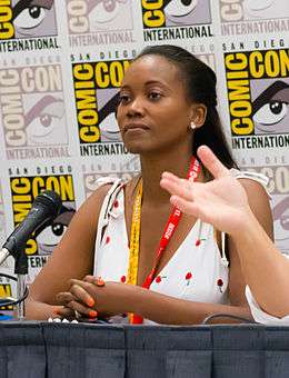 Photograph of a young African-American woman looking attentive; her long, black hair is pulled back into a low ponytail and she is wearing a sleeveless white top.