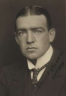  Man with hair centre-parted, wearing high white collar with tie, and a dark jacket. His facial expression is serious
