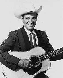 A man wearing a white cowboy hat and dark jacket, smiling broadly while holding a guitar