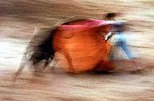 Blurred motion photo of bullfighter sweeping red cape over rushing bull.