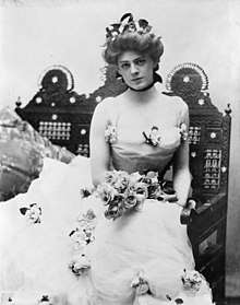 Ethel Barrymore, as a beautiful young woman, in a three-quarter length portrait, seated, facing front, wearing an elaborate gown and holding a bouquet of roses