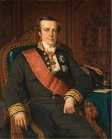 Portrait of a clean-shaven, square-faced man sitting in a chair. He wears a black suit with embroidered cuffs and a red sash over his right shoulder. Several awards are pinned to his coat.