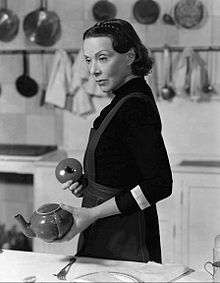 Black-and-white of a woman in a kitchen, holding a teapot.