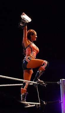A dark-haired woman standing on the turnbuckle of a wrestling ring, with one foot on the red ring ropes. She is wearing an orange crop top with dark shorts and dark-coloured wrestling boots. Her right hand is holding a wrestling championship in the air.