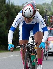 A woman, wearing a white and blue racing suit, grimaces as she races a bicycle on a paved road.
