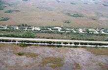 A color photograph taken from the air showing the Everglades bisected by a highway; at the bottom is a sawgrass field flooded with water bordered by a full canal; at the top are some homes and a dry sawgrass field