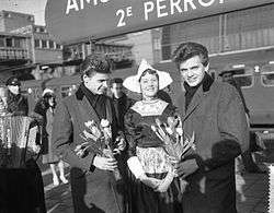 Two young men wearing long coats, holding bunches of flowers, standing either side of a woman in traditional Dutch clothing