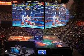 The V-shaped stage in a large events center surrounded by filled stands. Two people are sitting at a desk at the center of the stage, with two staffmembers standing next to them. Above the stage hangs a large cube with a monitor on each side, showing the Street Fighter match currently being played. Two presenters can be seen in the notch of the stage.
