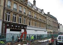 A row of 4- and 5-storey terraced buildings, with their ground floors boarded up. The buildings at the centre of the photo have stone facades; the others have brick. Workers are digging up the road along the frontage of the buildings.