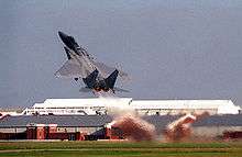 Gray jet fighter taking off at steep angle of attack, with full afterburner, as evident by hot gas ejected from its engines