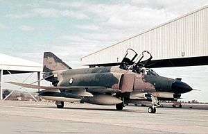 Two-seat military jet fighter painted in camouflage livery with open canopies, parked at airfield