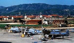  Three single-seat, piston-engined fighters under maintenance, with red-roofed building and a mountain range in the background