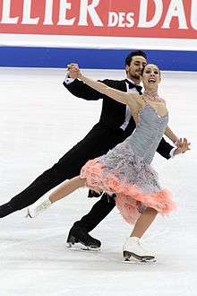 A man and woman ice dancing; the man, on the left, is wearing a classic tuxedo with a white-ruffled shirt, and the woman is wearing a knee-length light blue dress with a ruffled pink hem