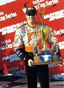 A man in his late twenties is wearing sunglasses along with orange racing overalls and is holding a trophy with both his hands.