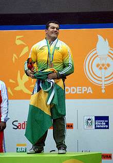 Brazilian athlete on the podium during the awards ceremony of the weightlifting competitions. Wearing the official uniform of the Brazilian delegation, he holds the flag of Brazil and a flowers bouquet. The bronze medal was hung around his neck.