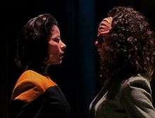 Two women are staring at each other while standing in a dark room. The woman on the right has ridges on her forehead and curly hair, while the one on the left has straighter hair and a uniform with a yellow strip.
