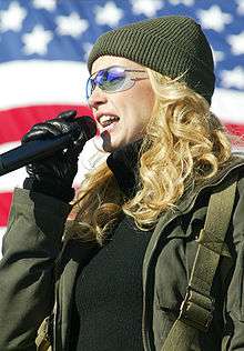 A blonde woman wearing a woollen hat and glasses singing into a microphone in front of the American flag
