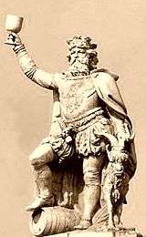 Gambrinus stands holding a chalice aloft, with his right foot atop a beer keg, and a goat to his left.
