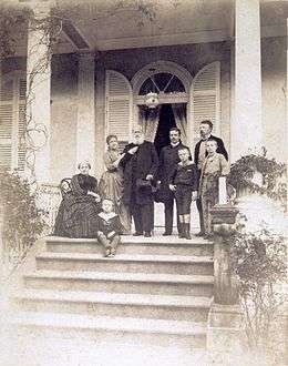 A group of people assembled on a columned porch at the top of a flight of steps, with one older woman seated, one younger woman leaning on the arm of an older bearded man, two younger men, and three small boys