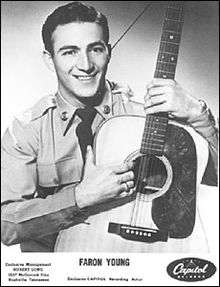 A dark-haired man wearing a shirt and tie, holding a guitar and smiling broadly