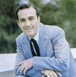 A dark-haired man wearing a blue jacket and black tie, smiling broadly