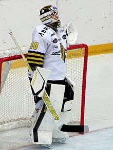 An ice hockey player standing in the goals with his body turned to the right of the camera. He is wearing a black and white uniform.