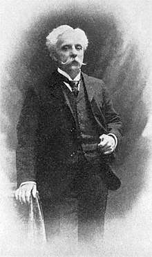 Portrait photograph of a standing man with white hair and large white moustache, wearing a dark three-piece suit