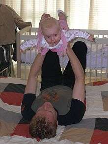 A young father lies on his back on a quilt on the floor. He holds his baby daughter up above him with his arms straight and his hands round her ribcage. The baby has her arms and legs stretched out and arches her back smiling directly at the camera.