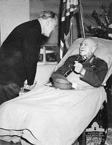 A man lies in a hospital bed, incongruously wearing an Army uniform instead of pyjamas. His peaked cap is on the blanket and he holds a baton in his hand. A man in a dark suit and pinstripe trousers bends over to talk to him. In the background are flowers, and a flag.
