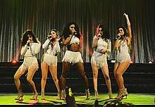 Five young women wearing white outfits consisting of short skirts and body-hugging tops are standing on a stage.