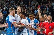 A distraught-looking man wearing a white jersey and shorts is seen on the left amid a group of other men, some similarly dressed while others wear blue jerseys and shorts, some of whom are touching him gently on the torso. At right are two men in red; one is holding a red card aloft in his right hand. In the background is a crowd. The scene is artificially lit.