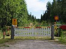 A simple white fence with a red and yellow gate behind it set across a dirt path in a green forest.