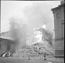 An apartment building is on fire and has partly collapsed in Central Helsinki after Soviet aerial bombing on 30 November 1939. A woman in a coat and a hat is passing by on the right next to the rubble and a car is on fire on the left.