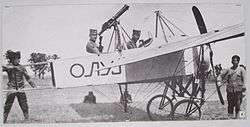 Two men seated in a World War I-era biplane, surrounded by technical personnel.