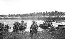 Soldiers walk through long grass. Other soldiers are arriving in landing craft in the lagoon behind them. In the background is a coconut plantation. The sky is overcast.