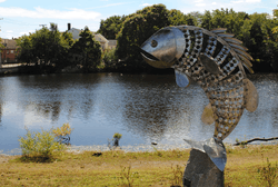 A sculpture of a fish in a park on the banks of a river.