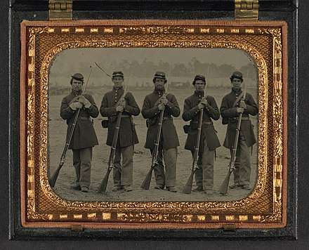 A sepia toned photograph of five soldiers standing at parade rest in a neat line