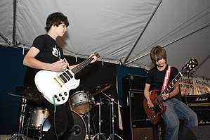 Two teenage guitarists are on stage. The one at left holds his guitar pick in his right hand, which is resting on it without playing. He stares over to the other one, who is crouching while playing. A drum kit is visible behind them.